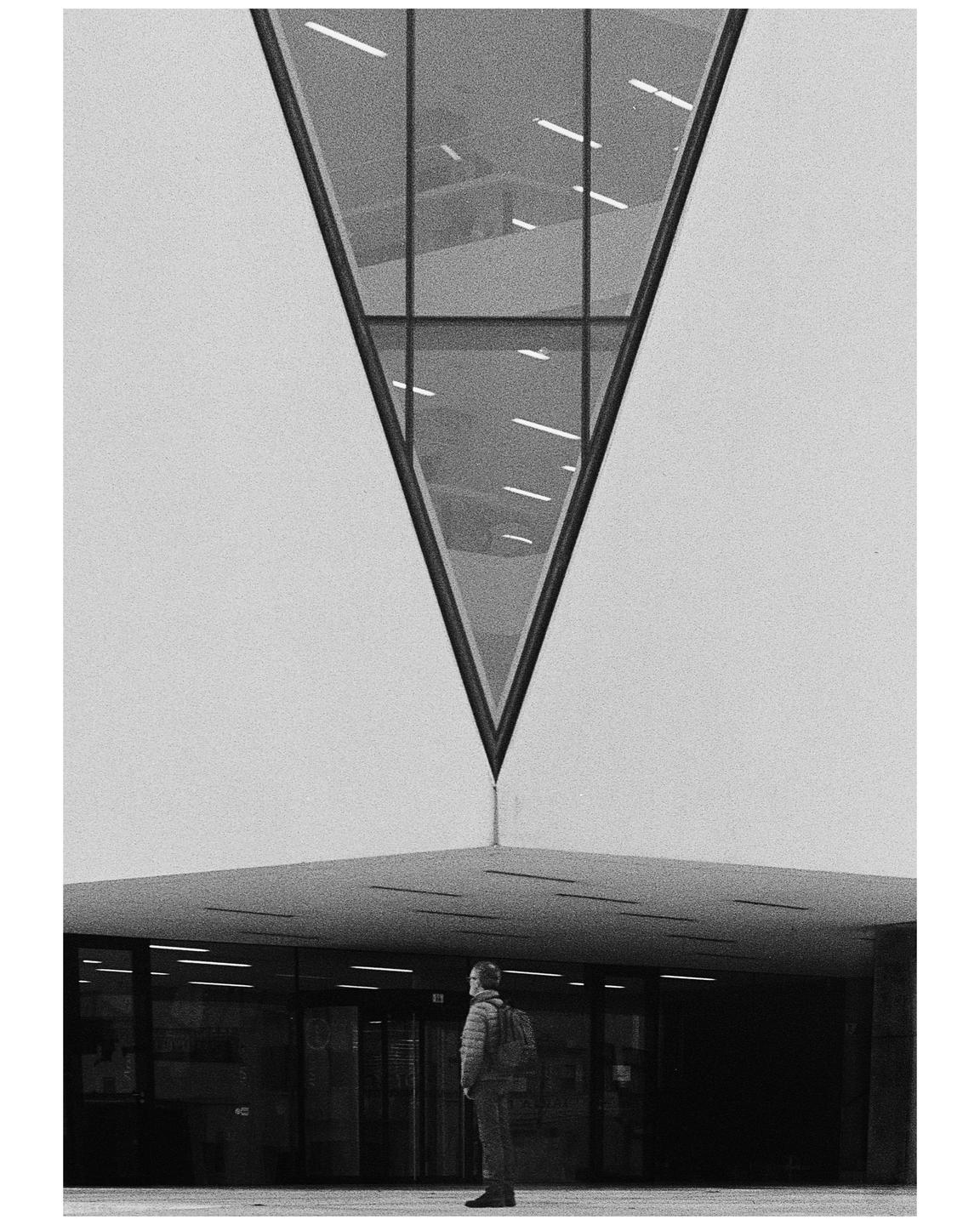 A man stands in front of a building with a large triangular window above him.