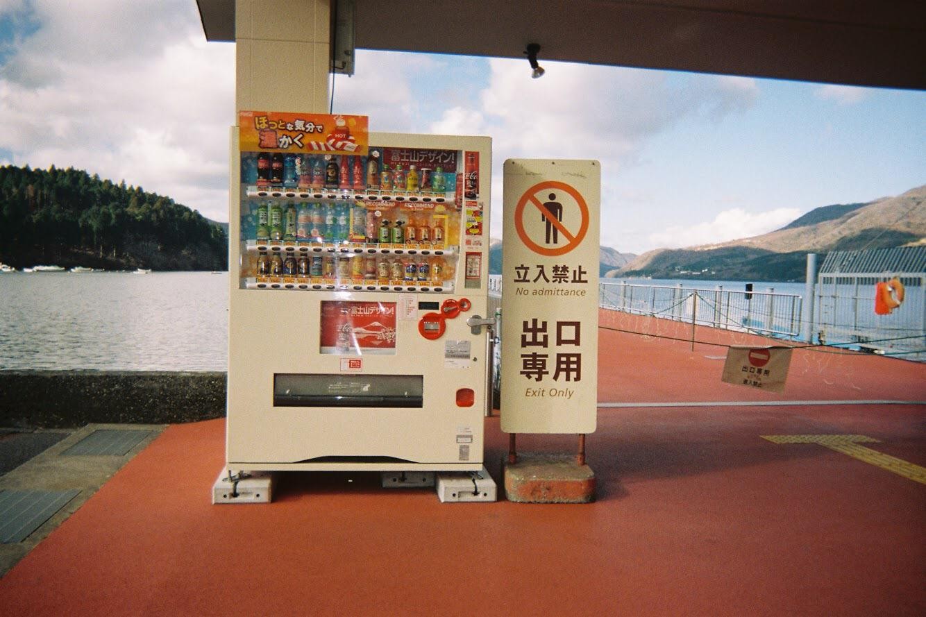 A vending machine and a "No Admittance" sign stand on a red platform next to a body of water with forested hills in the background.
