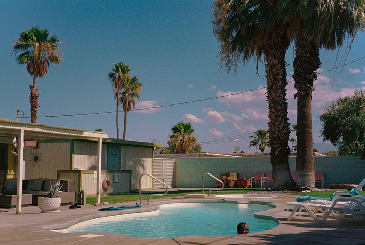 A man is in a swimming pool surrounded by palm trees and a building with a patio.