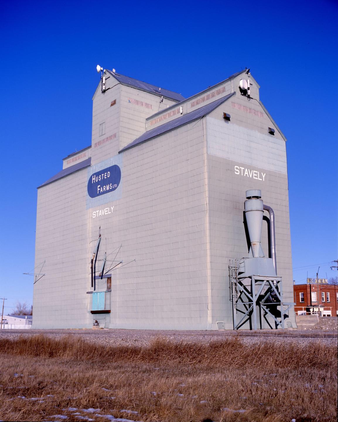 A large grain elevator with "Husted Farms Ltd" and "Stavely" written on its side stands under a clear blue sky.
