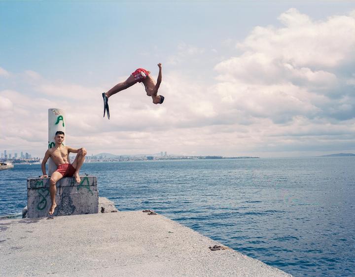 A man in red shorts sits on a concrete pier while another man in red shorts and flippers does a backflip over the water with a city skyline in the distance.