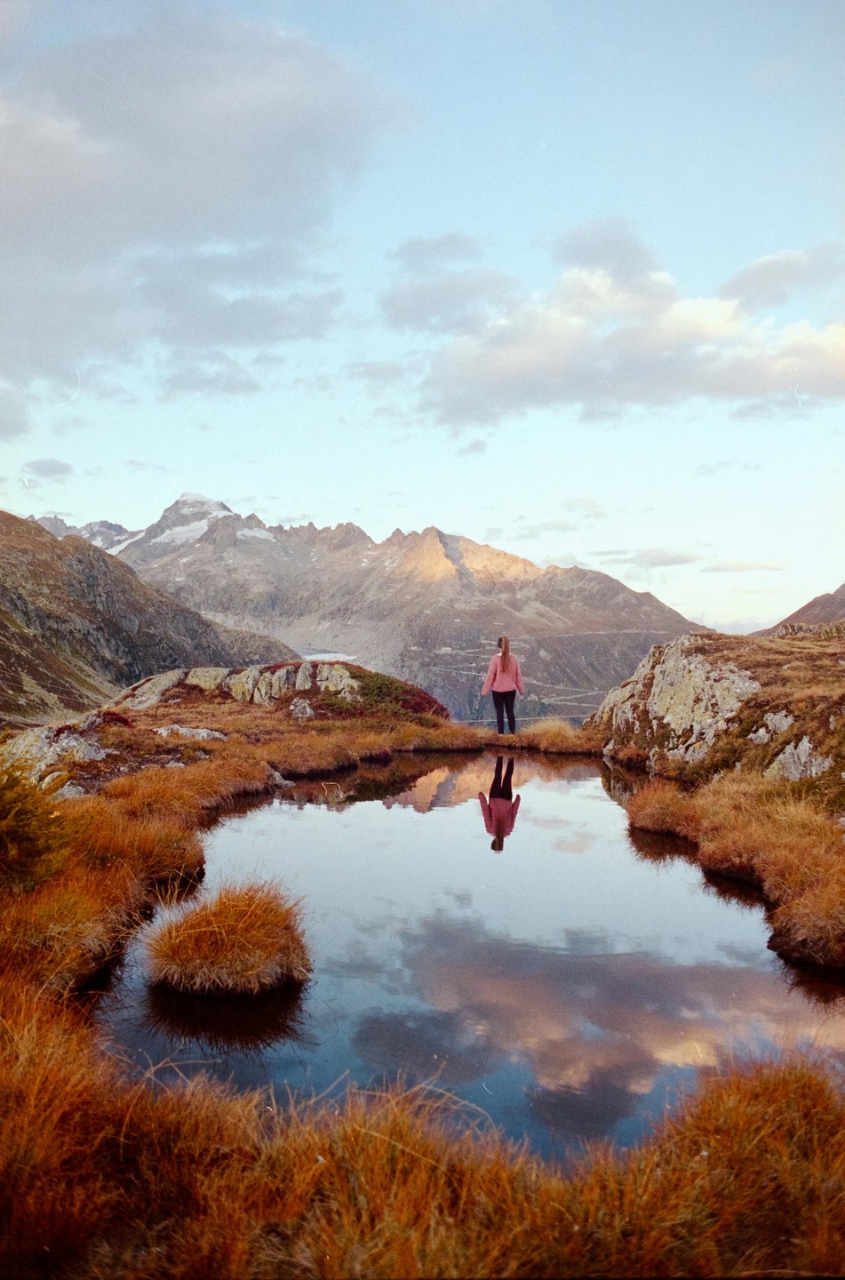 A woman stands by a small mountain lake reflecting the sky and surrounding peaks.