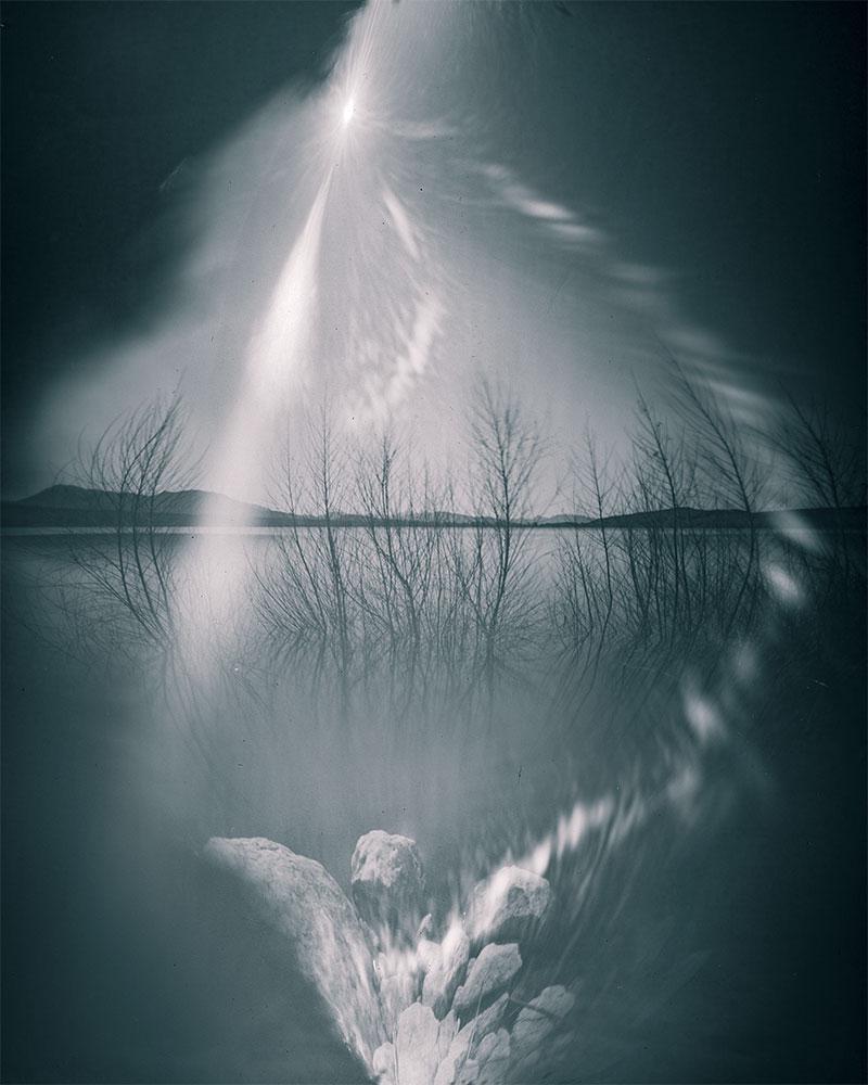 A long exposure photograph shows a lake with bare trees and mountains in the background, with rocks in the foreground and a bright, star-like light source in the sky.