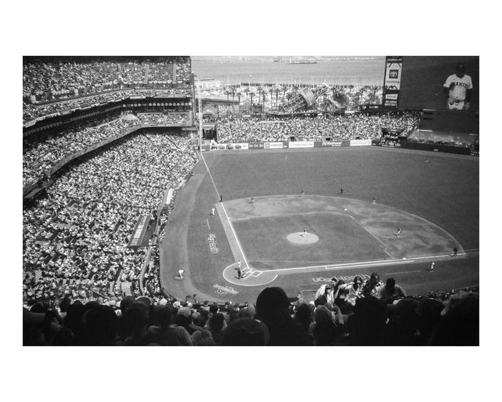 A baseball game is in progress at a stadium filled with spectators, with a body of water and city visible in the distance.