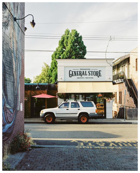 A white SUV is parked in front of the Fernwood General Store, which has a cream-colored awning and a sign that reads "Groceries + Provisions."