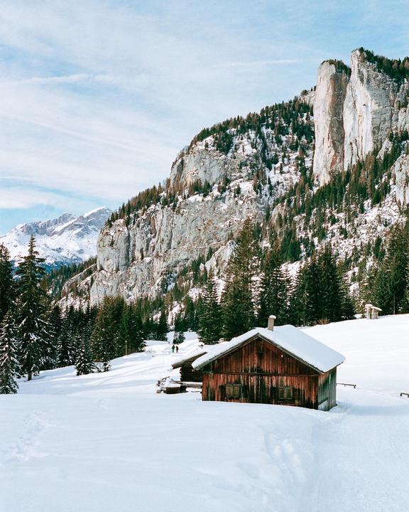 A wooden cabin sits in a snow-covered landscape with a mountain and forest in the background.