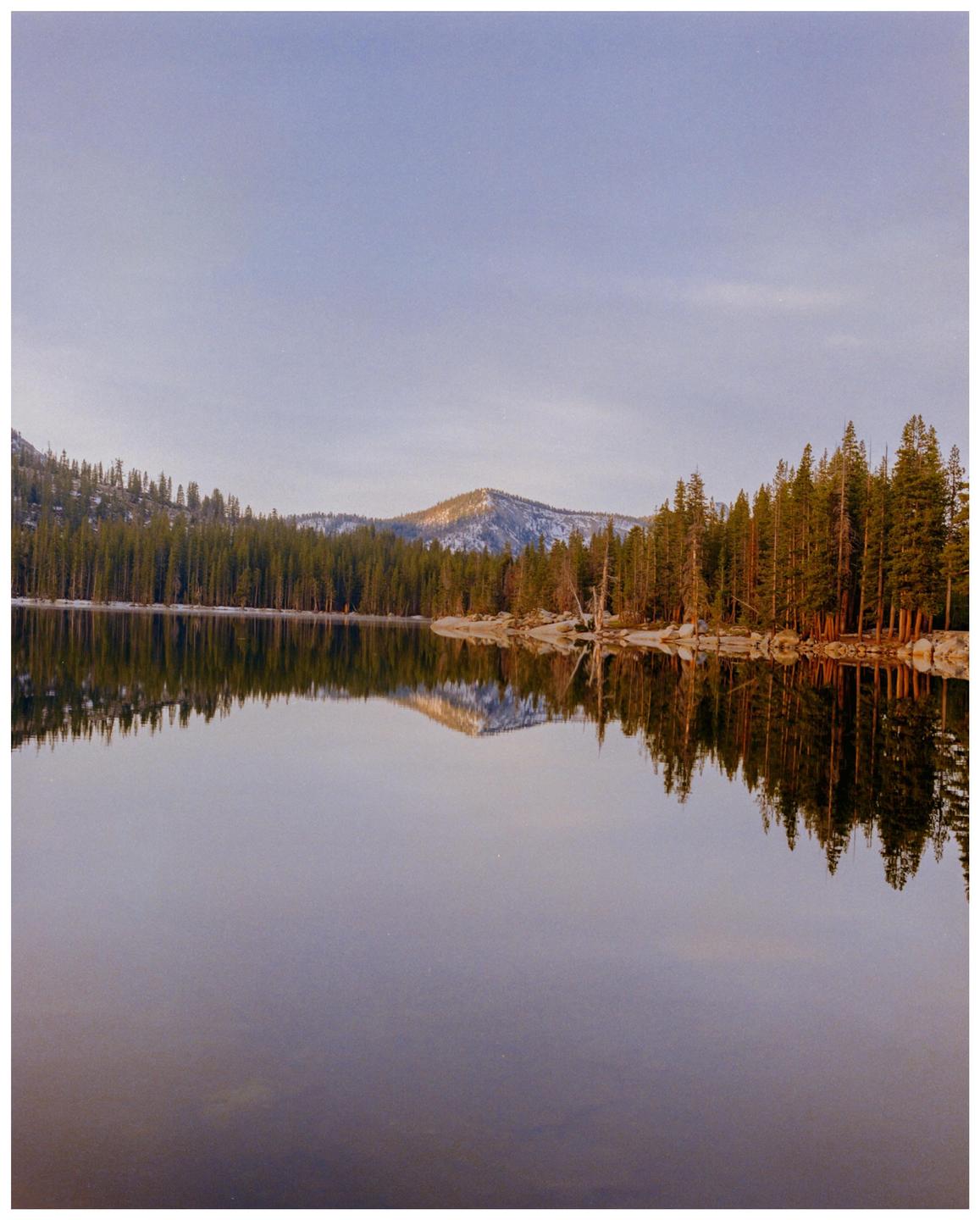 A calm lake reflects a forest and a snow-capped mountain under a clear sky.