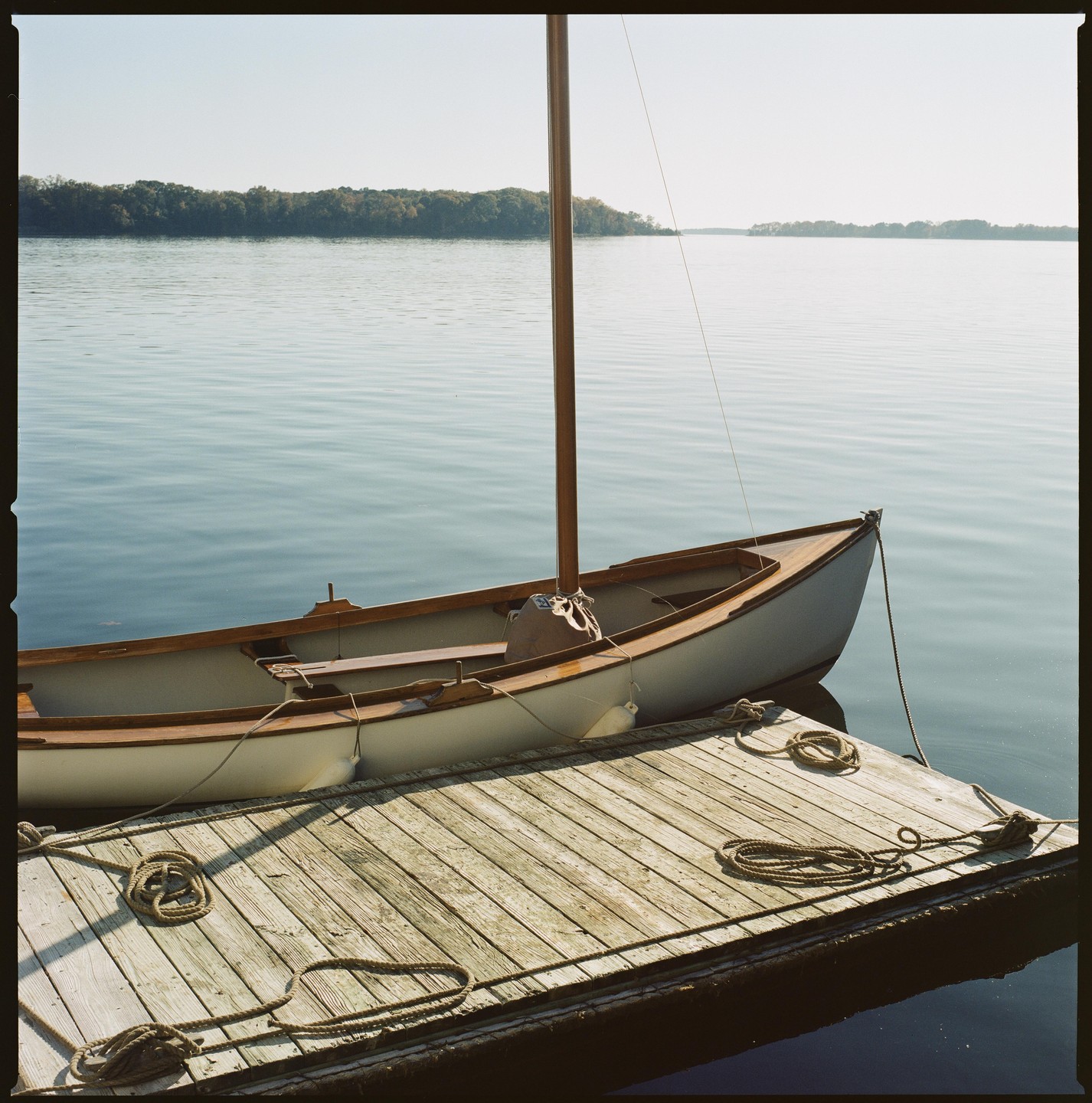 A small sailboat is docked next to a wooden pier on a calm body of water with a distant treeline.