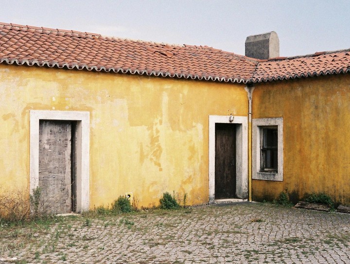 A yellow house with a red tile roof has two doors and one window.