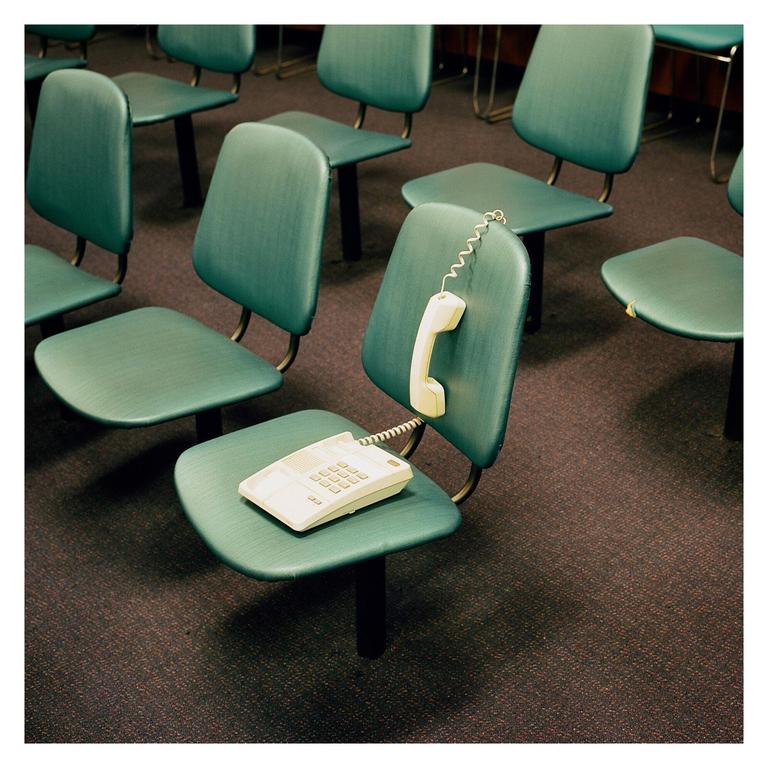 A white telephone with its receiver off the hook rests on a green chair in a room full of similar chairs.