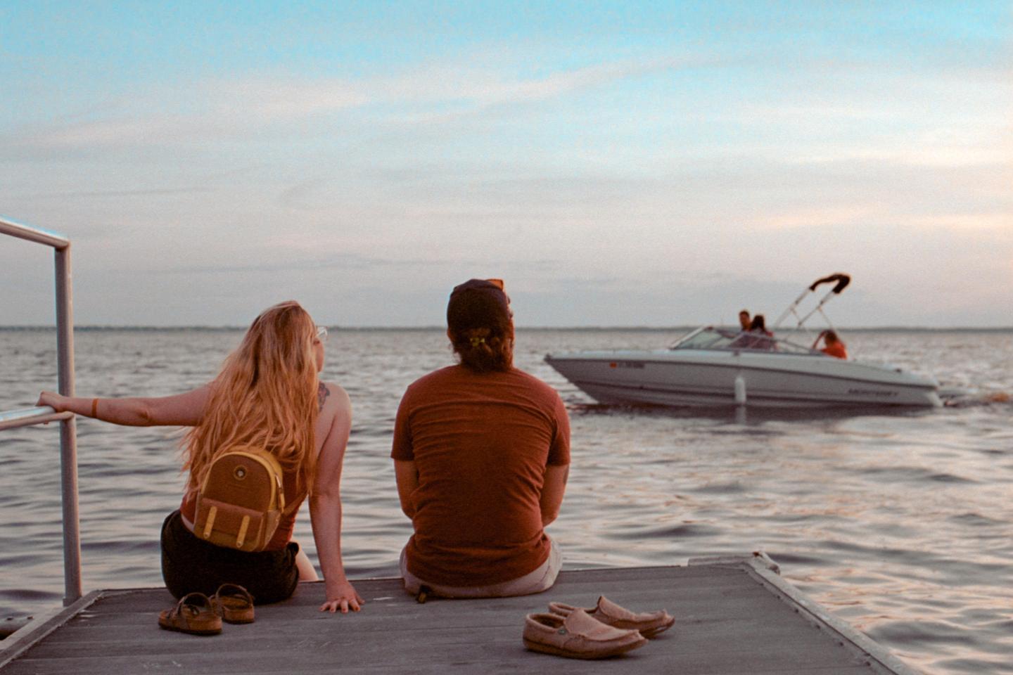 A woman and a man sit on a dock looking out at a boat on the water.