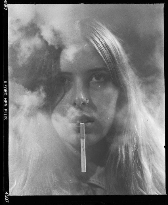 A woman with long hair holds a cigarette in her mouth as smoke billows around her face.