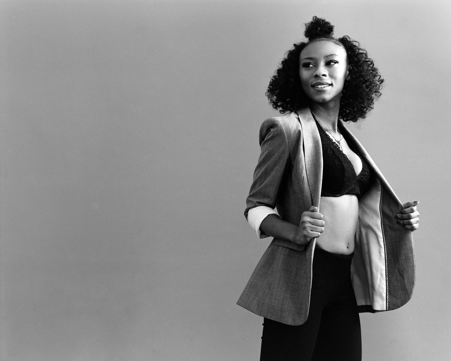 A woman in a blazer and bra smiles while looking to the side.