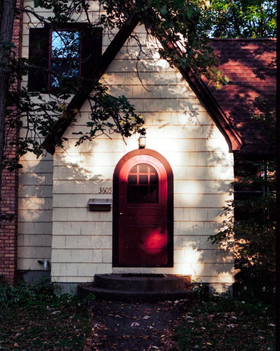 Sunlight illuminates the front of a house with a red door and a mailbox.