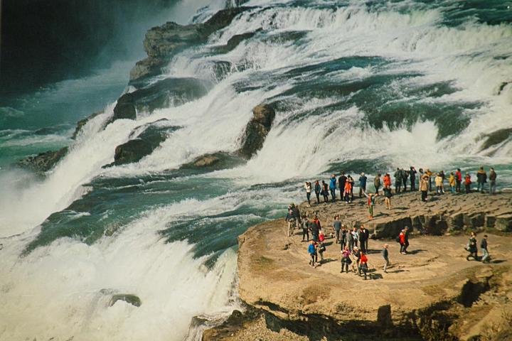 A large group of people stand on a rocky outcrop next to a powerful waterfall.
