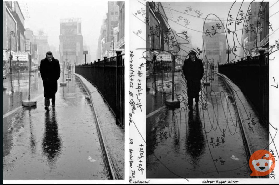 A man in a long coat walks on a wet street next to a fence with tall buildings in the background.