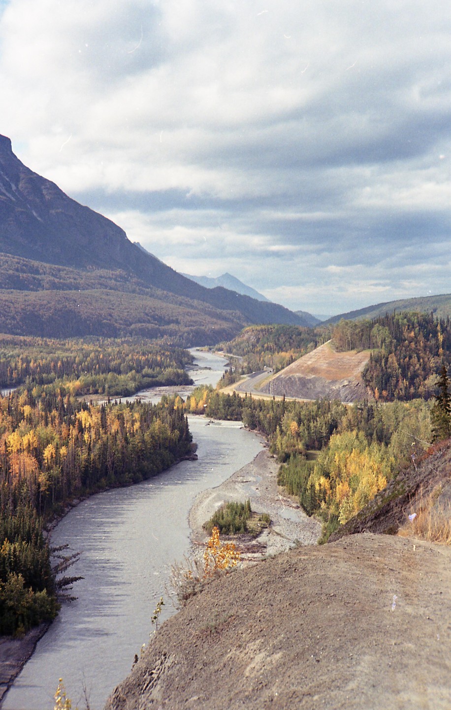 A winding river flows through a valley surrounded by mountains and autumn trees under a cloudy sky.