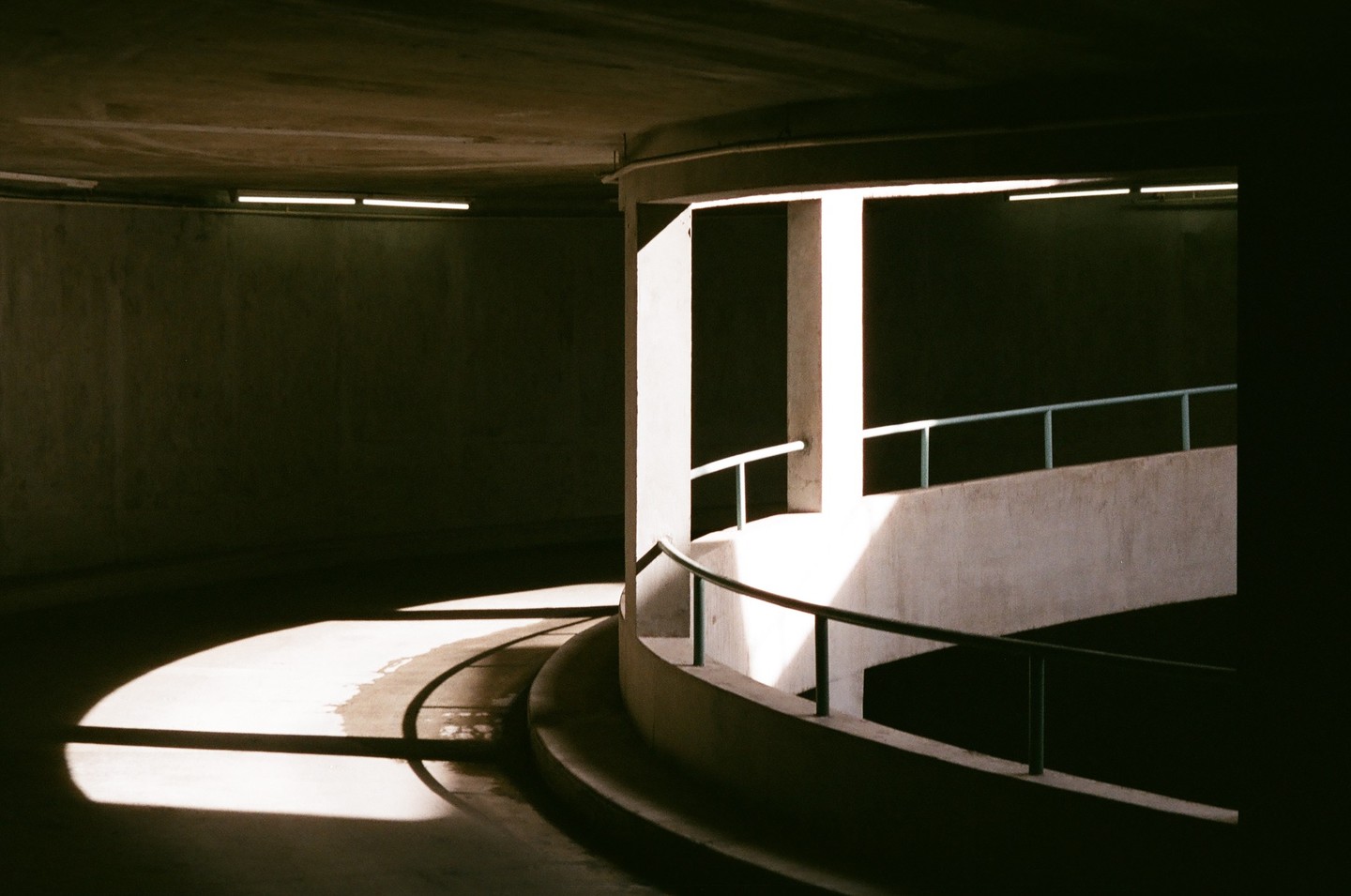 Sunlight casts strong shadows on a concrete parking garage ramp with railings.