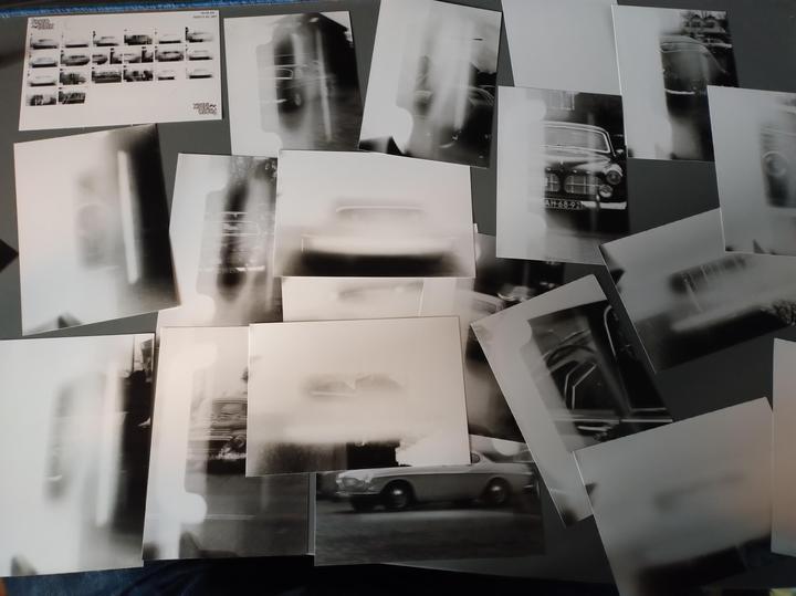 A collection of black and white prints of cars are scattered across a surface, with one contact sheet visible in the upper left corner.