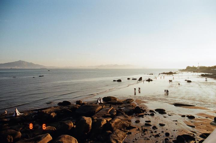 A wide beach with many people and large rocks is visible at golden hour, with mountains in the distance.