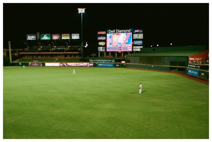 A baseball field is illuminated at night with a scoreboard and advertisements visible in the background.