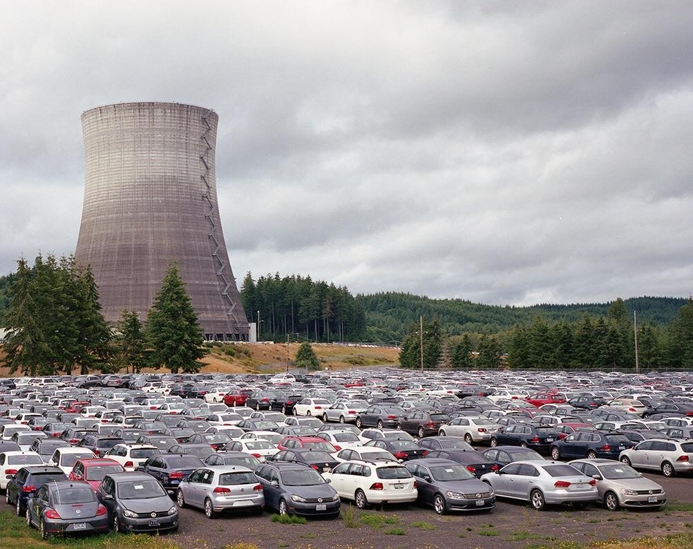 A large cooling tower stands behind a vast parking lot filled with cars, with a forest and cloudy sky in the background.