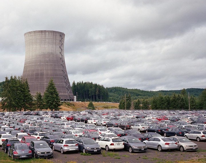 A large cooling tower stands behind a vast parking lot filled with cars, with a forest and cloudy sky in the background.