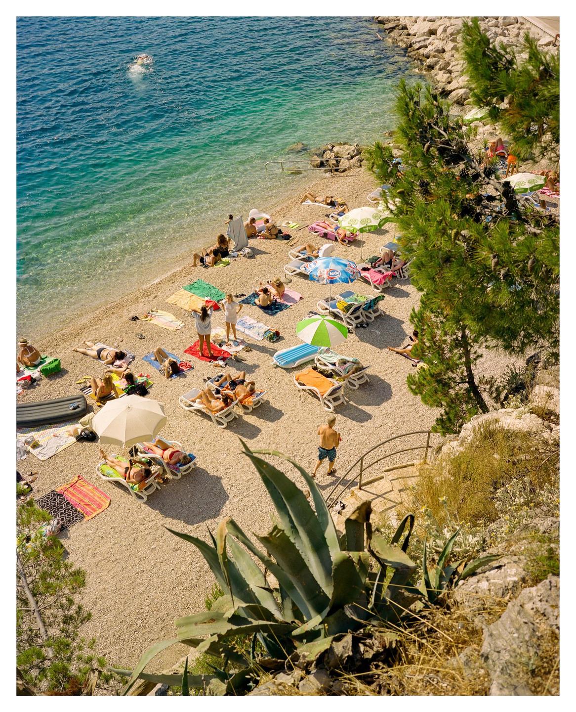 A high-angle view shows a crowded pebble beach with people sunbathing and swimming in the clear blue water.