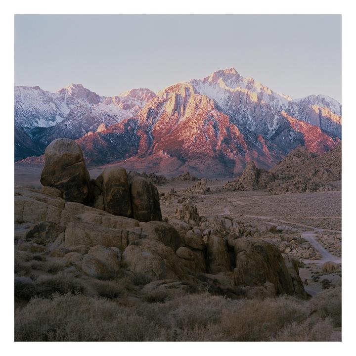 A mountain range with snow-capped peaks is illuminated by golden light behind a rocky, arid landscape.