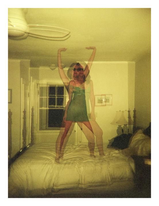 A woman in a green dress stands on a bed with her arms raised, captured with a double exposure effect.