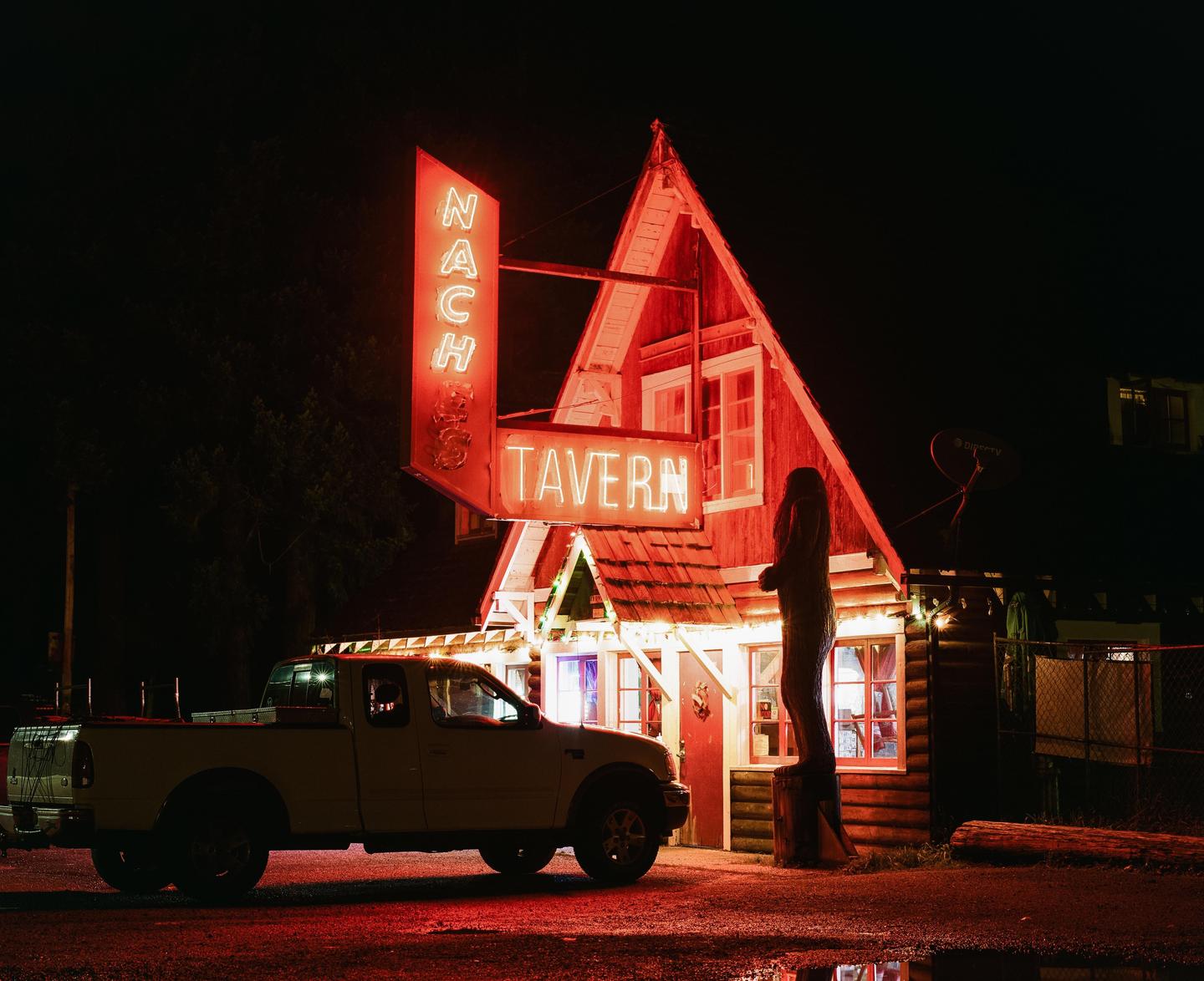 A white pickup truck is parked in front of a tavern with a neon sign that reads "Naches Tavern" at night.