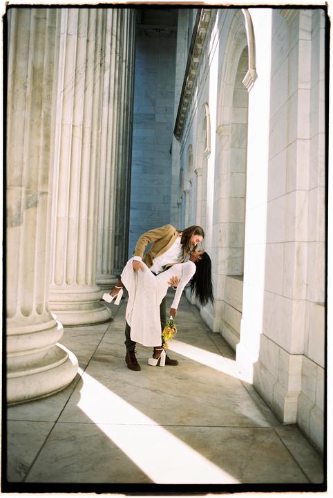 A couple dances in a hallway with large columns and arches, with sunlight casting shadows on the floor.
