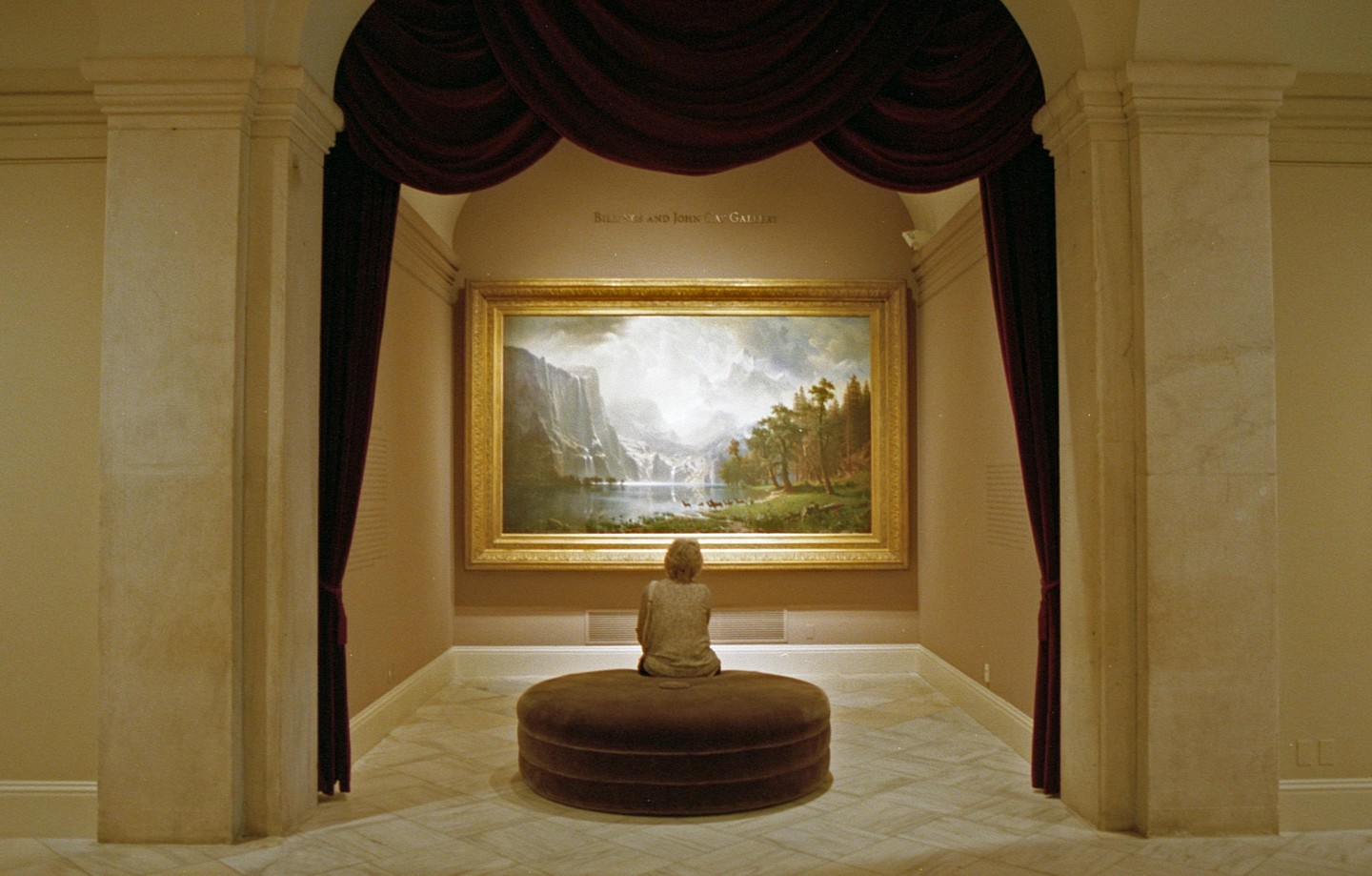 A person sits on a round ottoman, admiring a large landscape painting in a gallery.