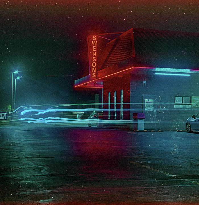 A building with a neon sign that reads "SWENSONS" is illuminated at night with light trails from vehicles in the foreground.