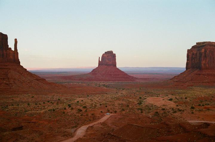 A vast desert landscape features towering rock formations under a pale sky with a dirt road leading towards a distant mesa.