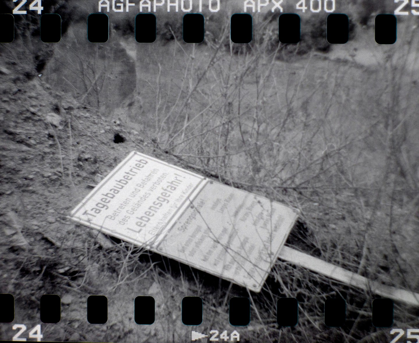 A warning sign with German text lies on the ground amidst dry brush and dirt.