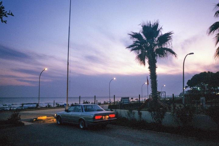 A car is parked on a road next to a beach with palm trees and streetlights at dusk.