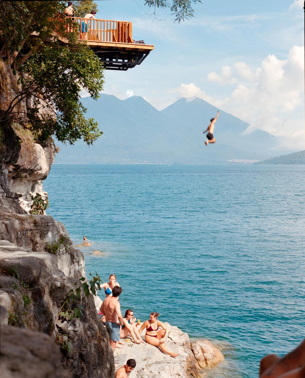 A man jumps from a wooden platform into a large body of water with mountains in the background while other people relax on rocks by the shore.