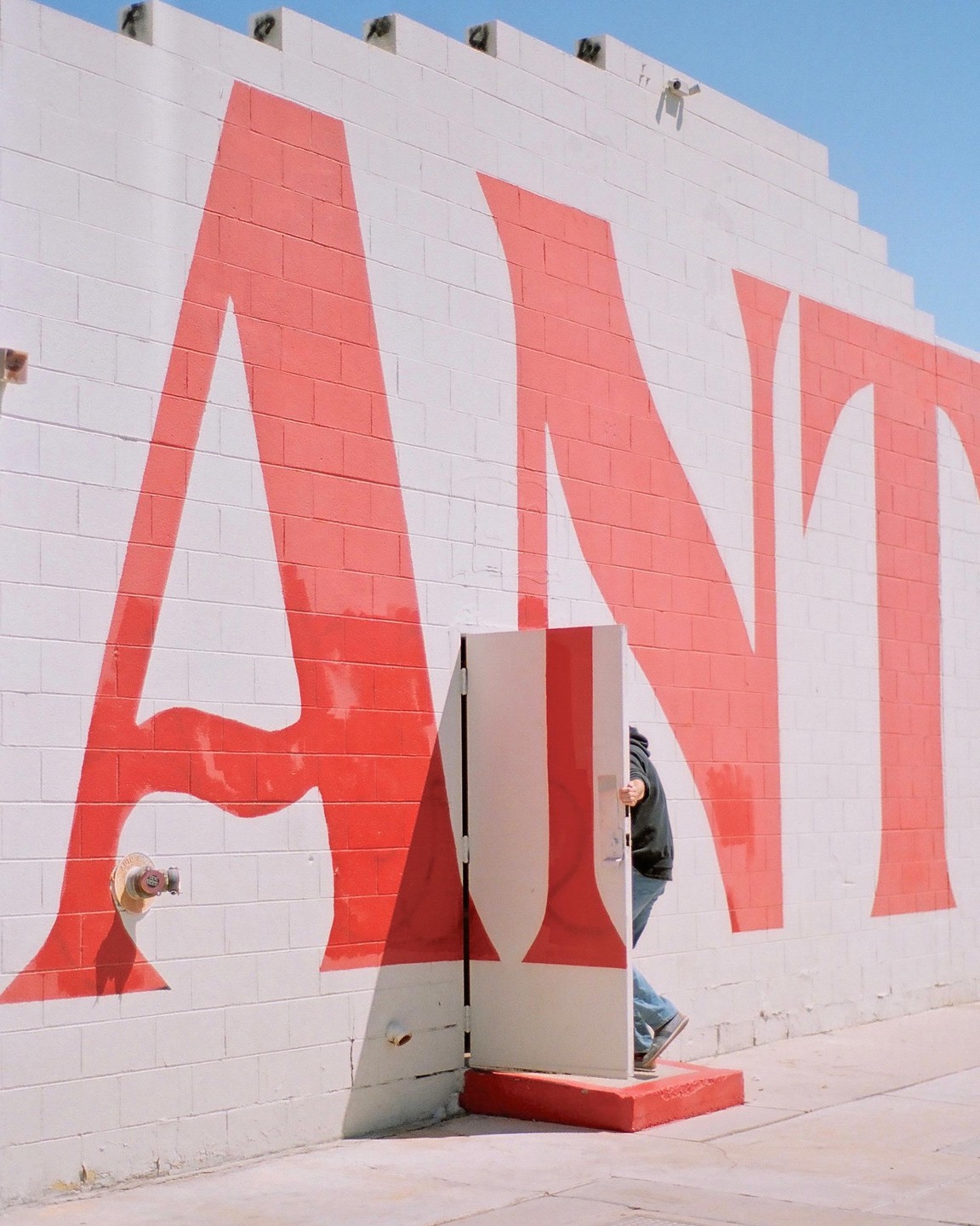 A person walks through a small door in a white brick wall with large red letters spelling "ANT" painted on it.