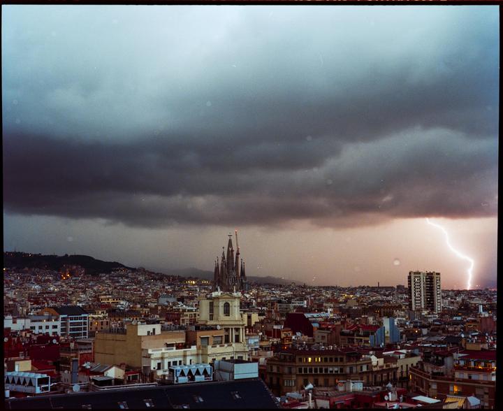 A lightning bolt strikes over a city skyline with a prominent cathedral under a stormy sky.