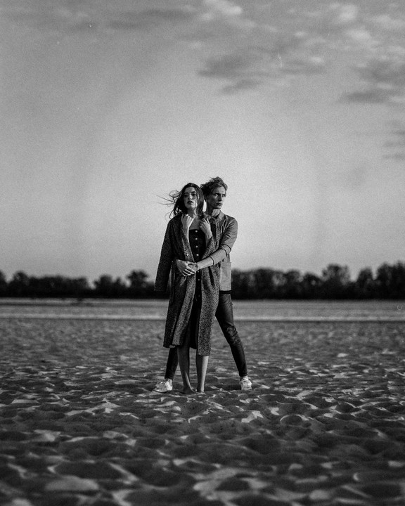 A couple stands on a sandy beach with trees and water in the background.