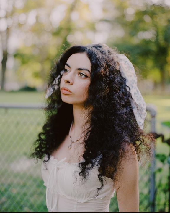 A woman with long, curly hair and a white top looks away from the camera in a garden.