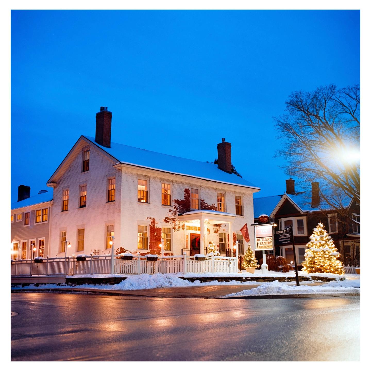 A snow-covered white building with illuminated windows and a porch is visible at dusk, with a Christmas tree and streetlights nearby.