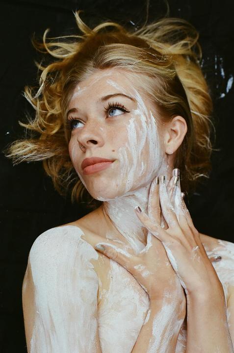 A woman with white paint on her face, neck, and shoulders looks up and to the right.