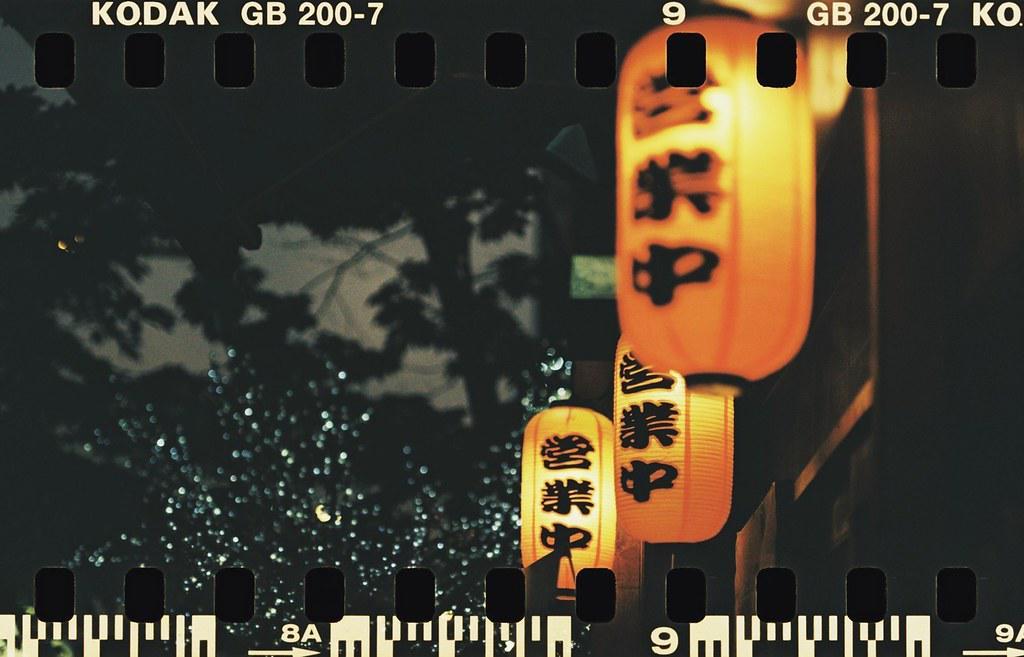 Three illuminated lanterns with Japanese characters hang on the right side of the frame, with a dark, out-of-focus background of trees and lights.