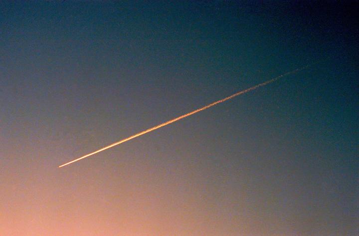 A single contrail streaks across a gradient sky from blue to orange.