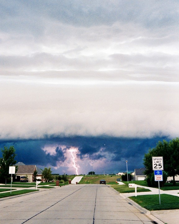A bolt of lightning strikes in the distance over a residential street with a speed limit sign in the foreground.
