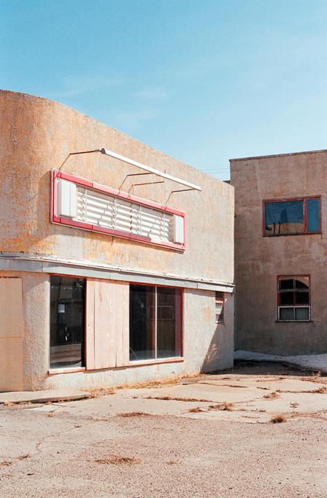 Two weathered buildings with a red-framed sign and boarded-up windows stand under a clear sky.
