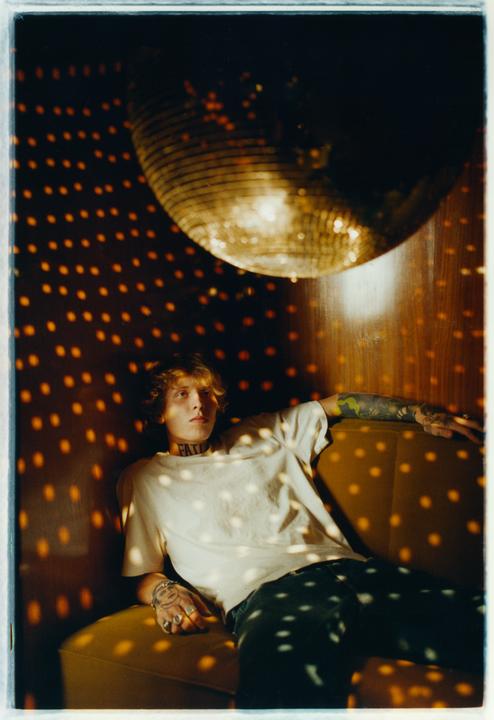 A man with tattoos on his arm and neck sits on a couch under a disco ball, which casts light spots on the wall and his body.