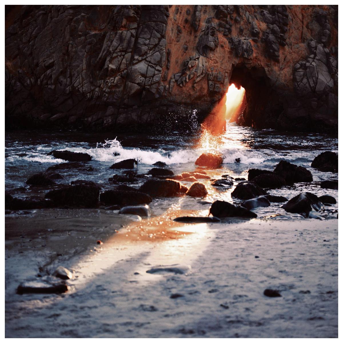 Sunlight streams through a rock arch onto a sandy beach with waves crashing over rocks.
